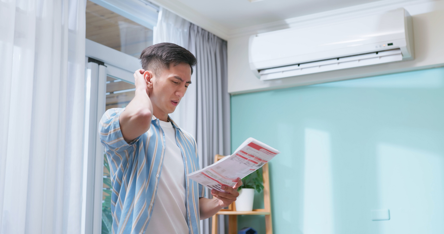 A man looks worried while reviewing a high summer electric bill beneath a running air conditioner.